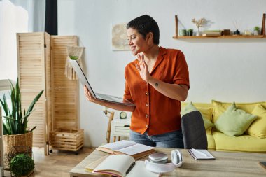 Neşeli öğrenci dizüstü bilgisayar kamerasına el sallıyor ve ders kitapları ve kulaklıklarla masanın yanında duruyor.