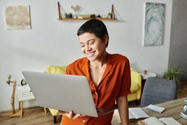 cheerful young woman with piercing in orange shirt looking joyfully at her laptop, education clipart