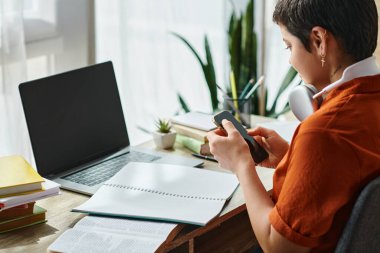 Kırmızı tişörtlü, kulaklıklı, ders kitabı ve telefonla çalışan kısa saçlı kadının yan görüntüsü.