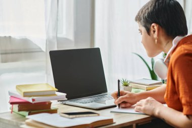 focus on laptop with blurred young student in front of it taking notes during online class clipart