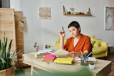 Günlük ev kıyafetleri içinde neşeli genç bir kadın masa başında çok çalışıyor ve kitap okuyor, evde eğitim görüyor.