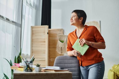 cheerful woman in casual attire standing next to desk with studying materials and looking away clipart