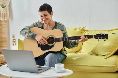 Neşeli bir kadın gitarla kanepede oturuyor online müzik dersine katılıyor, evde eğitim görüyor.