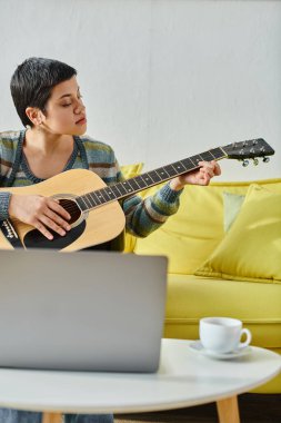 Genç, kısa saçlı bir kadının dizüstü bilgisayarın önünde oturup gitar çalmayı öğrendiği dikey çekim.