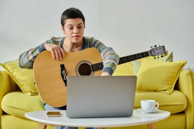 Online müzik dersine katılan genç bir kadın kanepede gitar çalıyor, evde eğitim görüyor.