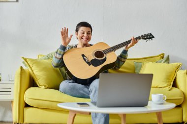 Çevrimiçi gitar dersine katılan neşeli genç bir kadın ve dizüstü bilgisayara el sallıyor, evde eğitim görüyor.