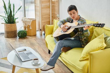 Evde gitar dersi alan, günlük ev elbiseli, kısa saçlı bir kadın.