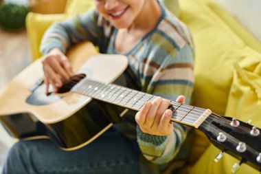 Gülümseyen, sıradan kıyafetli, uzaktan derste gitar çalmayı öğrenen genç bir kadın.