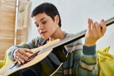 Uzaktan müzik dersinde gitar çalan, günlük kıyafetli, evde eğitim gören genç bir kadın.