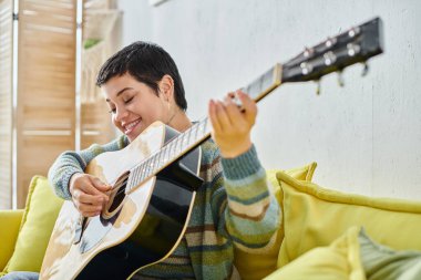 Uzaktan gitar dersinde kısa saçlı neşeli çekici kadın mutlu bir şekilde gülümsüyor, evde eğitim görüyor.