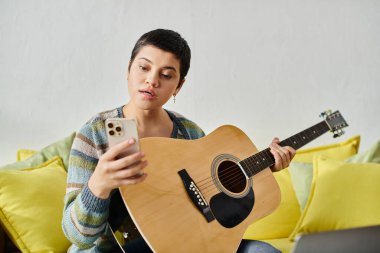Uzaktan müzik kursunda gitar çalan, günlük kıyafetli, kısa saçlı bir kadın.