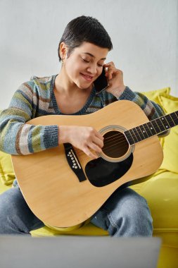 Online müzik dersi sırasında gitarla poz veren mutlu kadın telefonla konuşuyor.