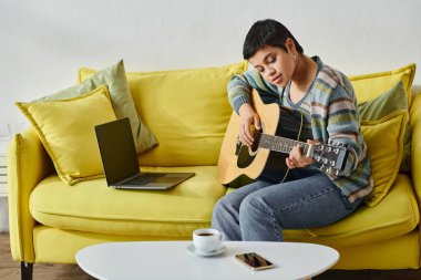 Kanepede dizüstü bilgisayarla oturup gitar çalmayı öğrenen genç bir kadın.