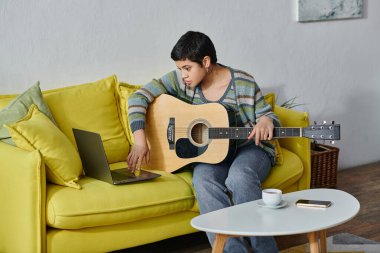 Çekici genç bir kadın elinde gitarla koltukta oturuyor ve dizüstü bilgisayara bakıyor, evde eğitim görüyor.