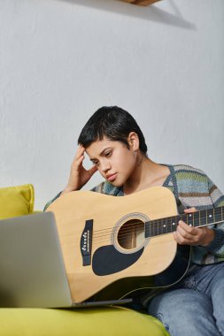 Sıradan giyinmiş, gitar çalmayı öğrenen, dizüstü bilgisayara ve eğitime bakan yorgun bir kadın.