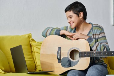 Neşeli kısa saçlı kadın online gitar dersi sırasında dizüstü bilgisayarına bakıyor, evde eğitim görüyor.