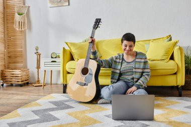 Uzaktan müzik dersinde gitar ve dizüstü bilgisayarla yerde oturan gündelik ev kıyafetleri içinde güzel bir kadın.