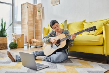 Online müzik dersinde gitar ve dizüstü bilgisayarı olan düşünceli genç bir kadın, evde eğitim görüyor.