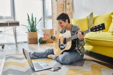 Online gitar dersi sırasında diz üstü bilgisayara gülümseyen kısa saçlı neşeli bir kadın.