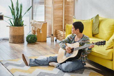 Neşeli bir kadın günlük kıyafetlerle yerde oturmuş gitar çalıyor ve başka tarafa bakıyor evde eğitim görüyor.