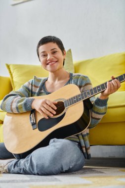 Kameraya gülümseyen gitarlı, evde eğitim gören neşeli bir kadının dikey görüntüsü.