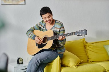 Kısa saçlı neşeli bir kadın gitar çalıyor ve mutlu bir şekilde gülümsüyor, evde eğitim görüyor.