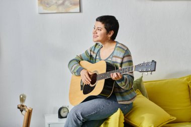 Güzel görünümlü esmer kadın elinde gitarla neşeyle gülümsüyor, evde eğitim görüyor.