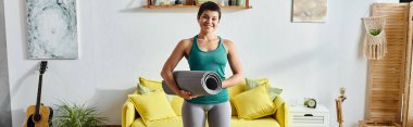 Spor kıyafetleri içinde neşeli bir kadın ayakta ve elinde spor minderi, spor ve pankart tutuyor.