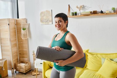 Kısa saçlı neşeli genç kadın fitness paspasıyla poz veriyor. Oturma odası arka planda, fitness...