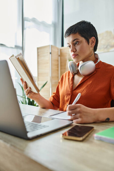 вертикальный снимок привлекательной женщины в оранжевой рубашке, делающий заметки и глядя на книгу, образование