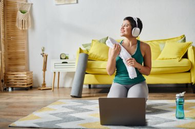 Spor kıyafetleri içinde neşeli bir kadın. Uzaktan kumanda fitness sınıfında su şişesi ve dizüstü bilgisayarın yanında oturuyor.