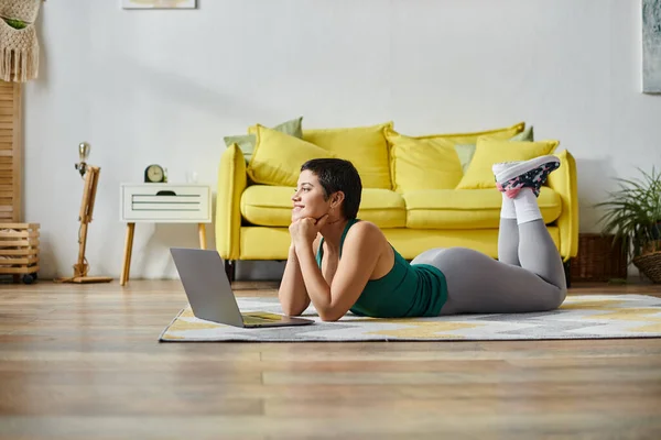 stock image jolly woman lying on floor during online fitness lesson smiling happily, hands under chin, fitness