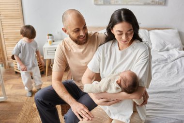 Bulanık oğulları arka planda oynarken yeni doğmuş bebeklerini kucağına alan modern sevgi dolu ebeveynlere odaklan.
