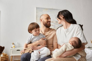 good looking parents holding their little son and newborn baby and smiling lovingly at each other clipart