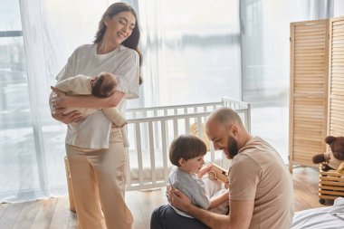 beautiful mother holding her baby boy and looking lovingly at her husband and son, family concept clipart