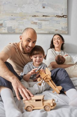 jolly bearded father hugging his son smiling at camera with his wife and newborn baby on backdrop clipart