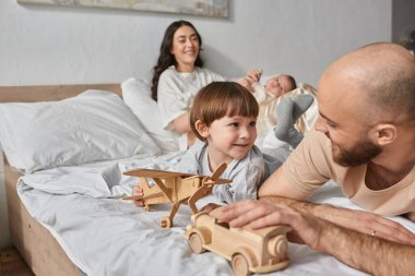Bulanık bir anneyle yatakta yatan baba ve küçük oğlana odaklan. Yeni doğmuş bebeği arka planda tut.