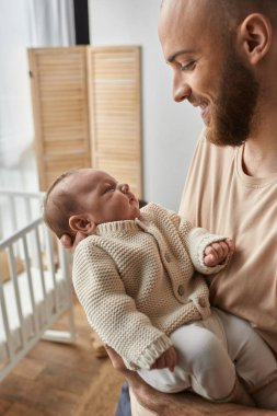 vertical shot oh jolly bearded man looking lovingly at his newborn baby boy while holding him clipart