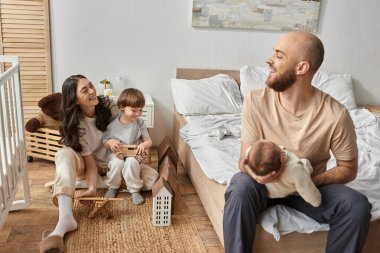 Neşeli modern ebeveynler çocuklarıyla yatak odalarında vakit geçirip birbirlerine gülümsüyorlar.
