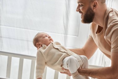 Ev elbiseli yakışıklı, neşeli baba yeni doğmuş bebeğini beşiğine koyuyor, aile kavramı.