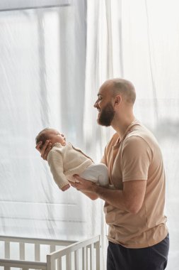 Yeni doğmuş oğlunu kucağında tutan ve ona sevgiyle gülümseyen mutlu bir baba resmi.