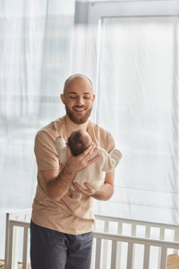 İyi görünümlü sakallı bir babanın bebeğini beşiğin yanında tutuşu, aile kavramı.