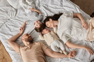 Dört kişilik ev elbiseli mutlu bir aile yatakta uzanıyor ve mutlu bir şekilde gülümsüyor, modern ebeveynlik.