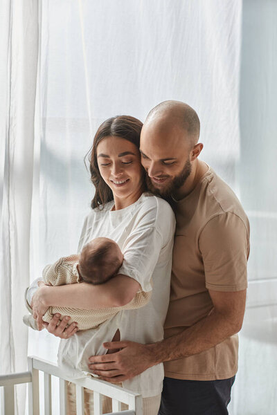 vertical shot of happy couple holding their newborn baby boy and smiling at him next to crib, family