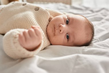 Bej hırkalı, yatakta yatıp kameraya bakan sevimli, yeni doğmuş bir bebeğin portresi.