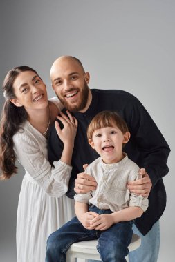 Uzun bir sandalyede oturan küçük oğluna sarılan neşeli modern ebeveynlerin dikey çekimi.