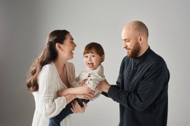 Neşeli anaokulu çocuğu, neşeli ailesinin ellerinde kameraya dilini uzatıyor.