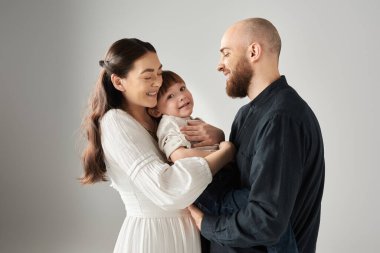 Beyaz elbiseli neşeli bir anne, küçük oğluna sarılırken kocası onlara bakıyor.