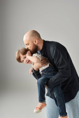 Mutlu sakallı babanın neşeli oğlunu tutarken ve öperken çekilen dikey fotoğrafı, modern ebeveynlik.