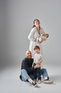 Mutlu bir anne yeni doğmuş bebeğini kucağında tutarken kocası yerde oturmuş küçük oğluna sarılıyor.